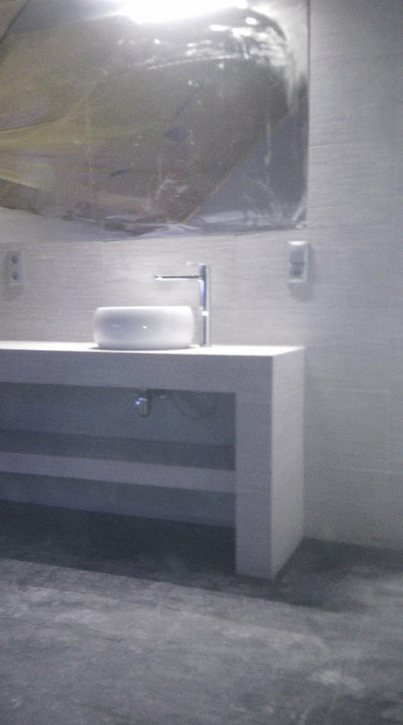 Nowoczesna łazienka: biała umywalka nablatowa z chromowaną baterią na blacie z szarymi pasami, lustro zabezpieczone folią, szare płytki na ścianach.