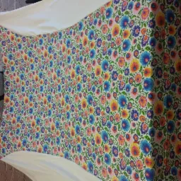 Kobieta prezentuje pościel w kolorowe kwiaty na tle ceglanej ściany i drewnianej podłogi. Widoczny fragment białej tkaniny i uśmiechnięta twarz kobiety.