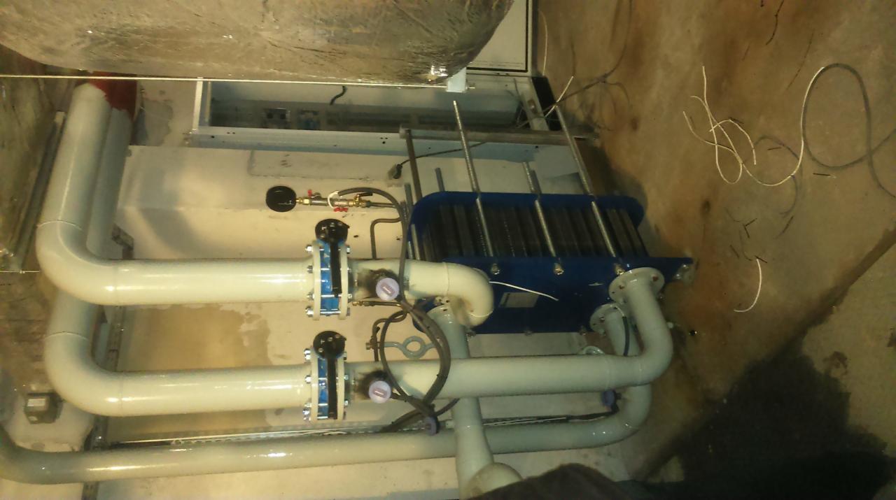 Wymiennik ciepła płytowy, błękitny, zamontowany w kotłowni, z rurami w kolorze kremowym, z manometrami, w surowym pomieszczeniu technicznym z betonową podłogą.