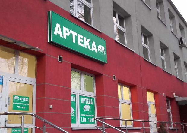 Fasada budynku apteki z dużym, zielonym szyldem z napisem 'Apteka' i logo sieci. Widoczne okna na dwóch kondygnacjach, budynek pomalowany na czerwono i szaro.