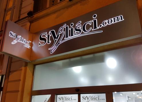 Podświetlany szyld z nazwą 'Stylisci.com' nad wejściem do salonu w wieczornym oświetleniu, widoczny z dołu pod kątem.