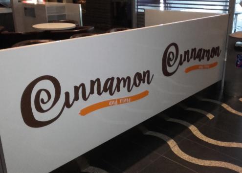 Biała przegroda z brązowym napisem 'Cinnamon and more' z pomarańczowym akcentem, widoczna w kawiarni lub restauracji.