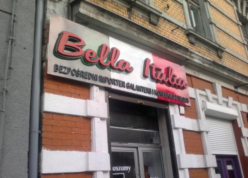 Szyld sklepu 'Bella Italia' z czerwono-zielonym napisem na tle włoskiej flagi, informujący o bezpośrednim imporcie galanterii i konfekcji z Włoch, umieszczony nad wejściem do lokalu z przeszklonymi...