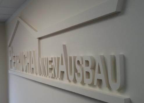 Białe, trójwymiarowe litery tworzące napis 'Pierucha Innenausbau' zamontowane na białej ścianie, podświetlone z góry, z perspektywy z dołu i z boku.