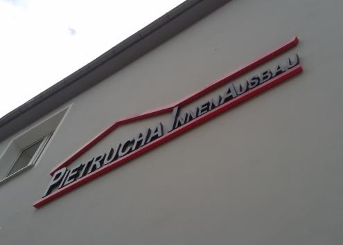 Zewnętrzny szyld firmy 'Pietrucha Innenausbau' z czarnymi literami na czerwonym tle, umieszczony na jasnej ścianie budynku z oknem po lewej stronie.