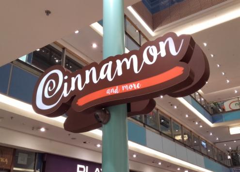 Brązowy, przestrzenny szyld 'Cinnamon and more' z białym napisem umieszczony na zielonym słupie wewnątrz galerii handlowej, widoczne oświetlenie sufitowe i fragmenty innych sklepów.
