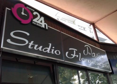 Szyld reklamowy 'Studio Pasji Florystyczne' z logo czynne 24h, zamontowany nad wejściem do lokalu, widoczny z dołu pod kątem.