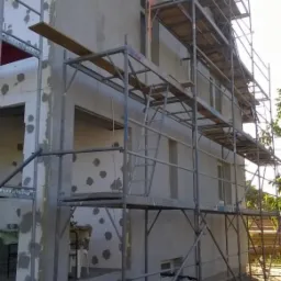 Budynek w trakcie ocieplania, widoczne rusztowania, częściowo otynkowana elewacja z szarym tynkiem, obok stoją rowery, widok z dołu, słoneczny dzień.