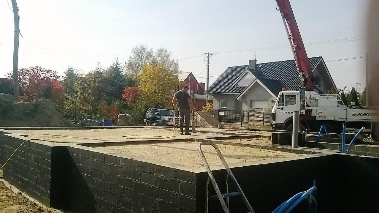 Fundamenty domu w trakcie izolacji, widoczny pracownik z wężem, betonowa pompa i otoczenie budowy w słoneczny dzień.