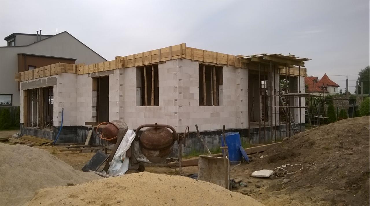 Budowa domu z betonu komórkowego w trakcie realizacji. Widoczne ściany, drewniane szalunki okienne i drzwiowe, oraz dwie betoniarki. W tle budynki mieszkalne.