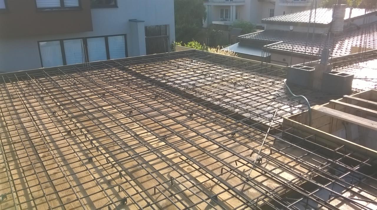 Zbrojenie stropu budynku jednorodzinnego z widoczną siatką zbrojeniową, deskowaniem i elementami szalunkowymi. W tle budynki mieszkalne.