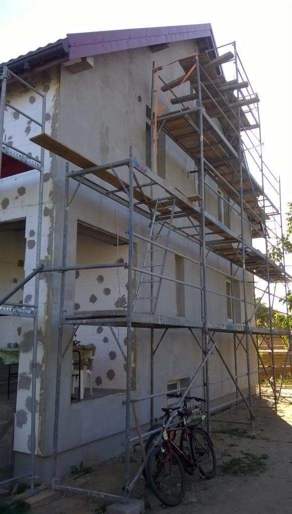 Budynek w trakcie ocieplania, widoczne rusztowania, częściowo otynkowana elewacja z szarym tynkiem, obok stoją rowery, widok z dołu, słoneczny dzień.