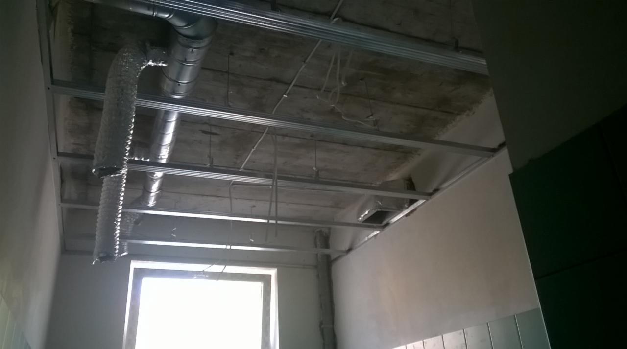 Montaż instalacji wentylacyjnej pod sufitem w surowym wnętrzu. Widoczne przewody wentylacyjne, metalowa konstrukcja podwieszana i okno w tle.