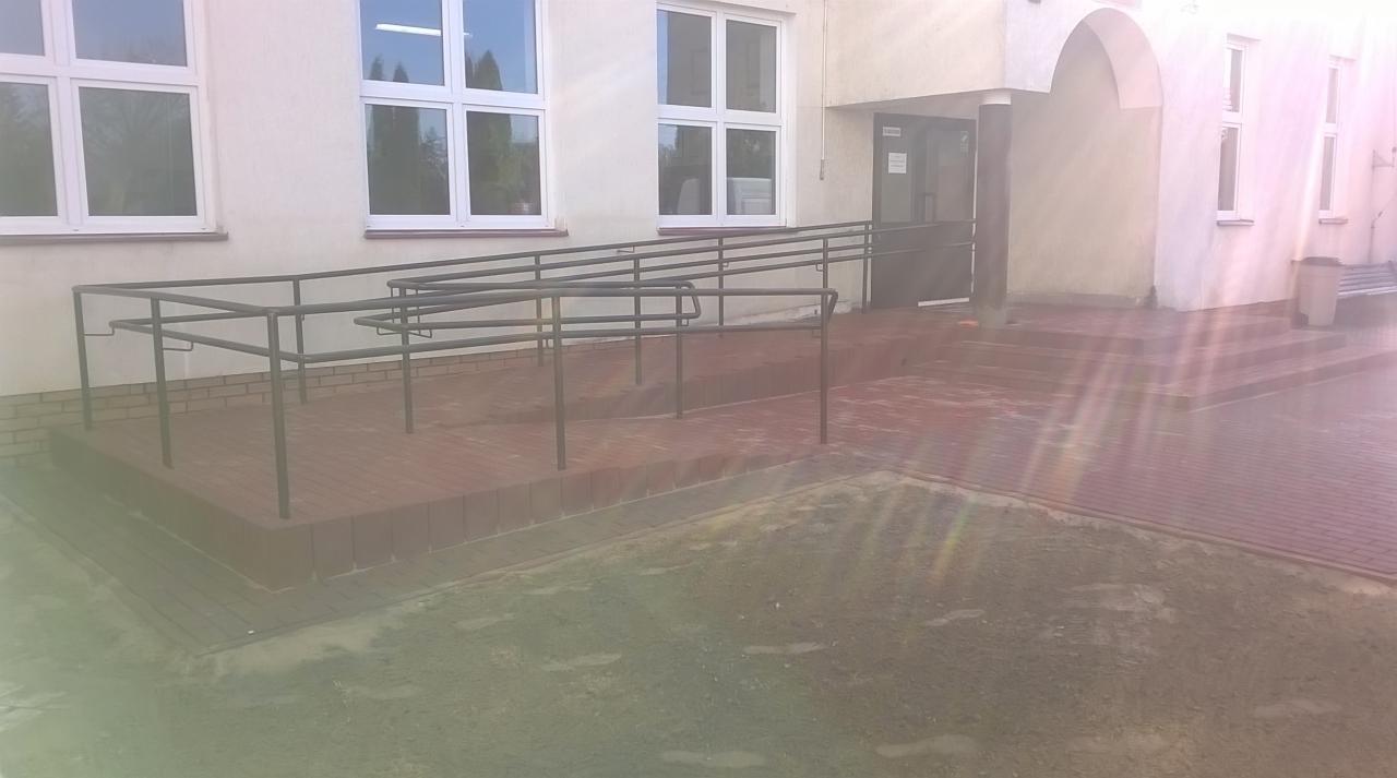 Rampa dla niepełnosprawnych z metalową barierką, wyłożona czerwoną kostką brukową, prowadząca do wejścia budynku z białymi oknami.