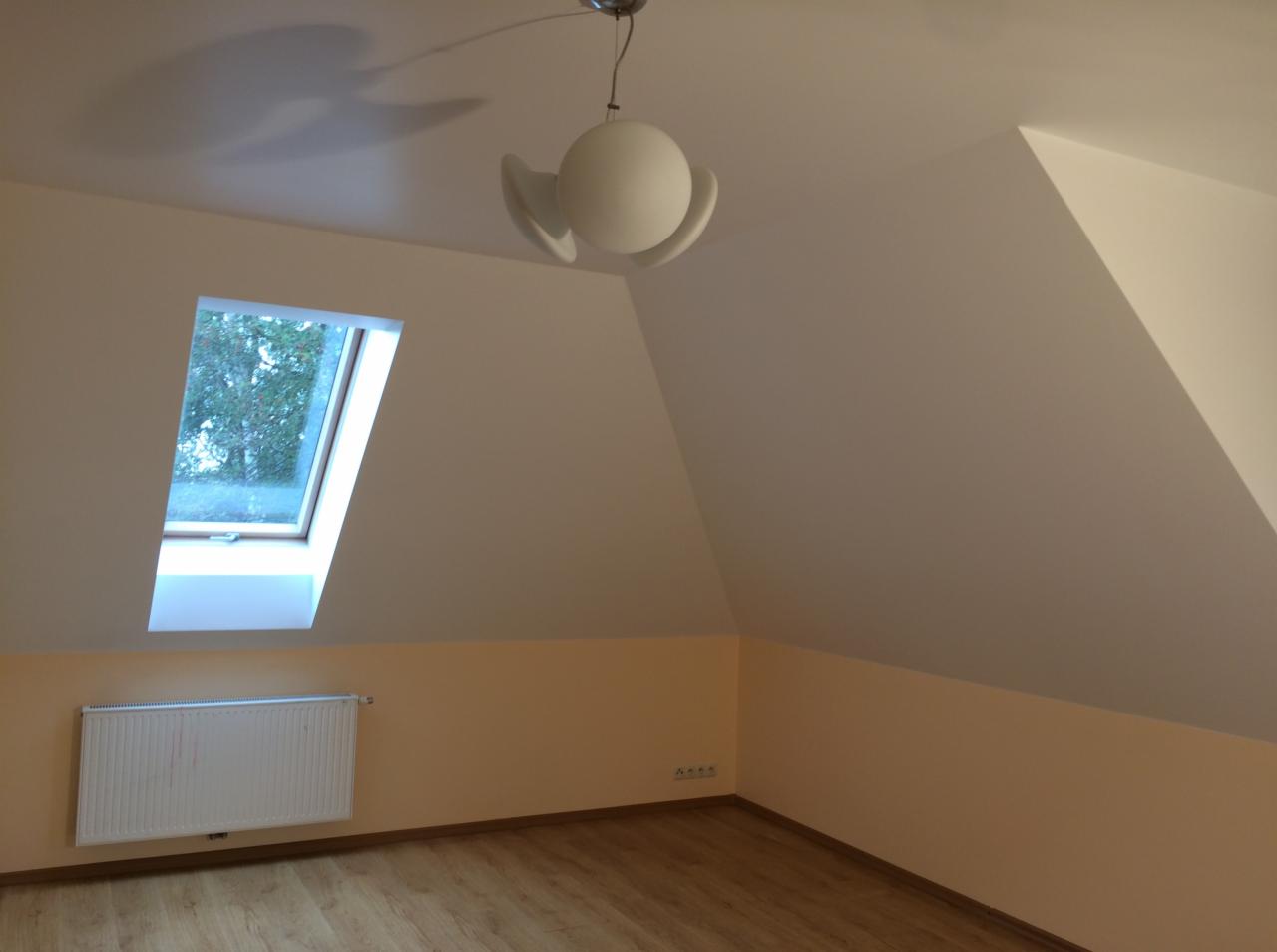 Poddasze z oknem dachowym, pomalowane ściany w kolorze ecru i białym, drewnopodobna podłoga, biały grzejnik pod oknem i nowoczesna lampa sufitowa.