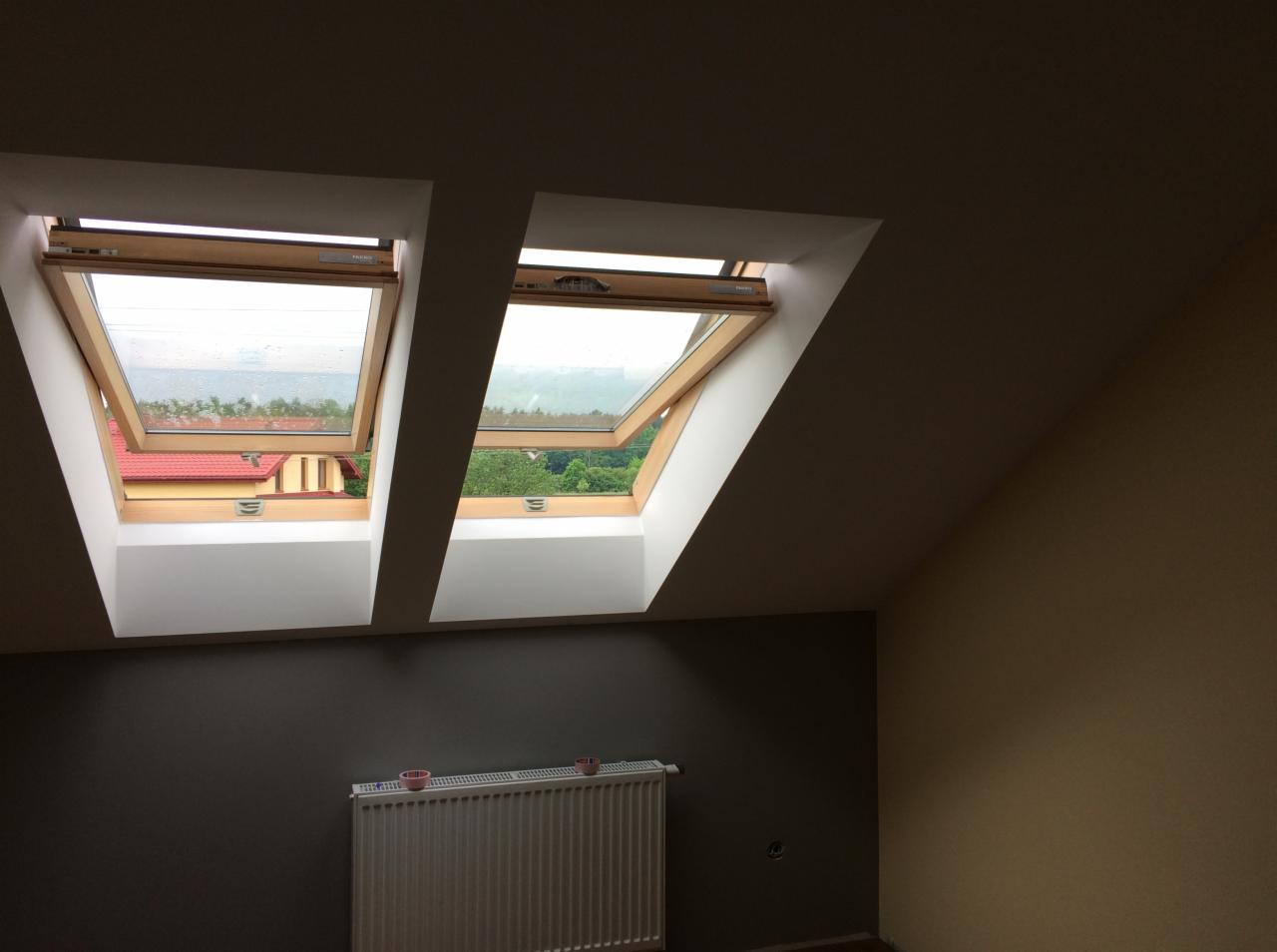 Poddasze po remoncie z dwoma otwartymi oknami dachowymi, widoczne fragmenty pomalowanych ścian w kolorze szarym i beżowym, biały grzejnik pod oknami.