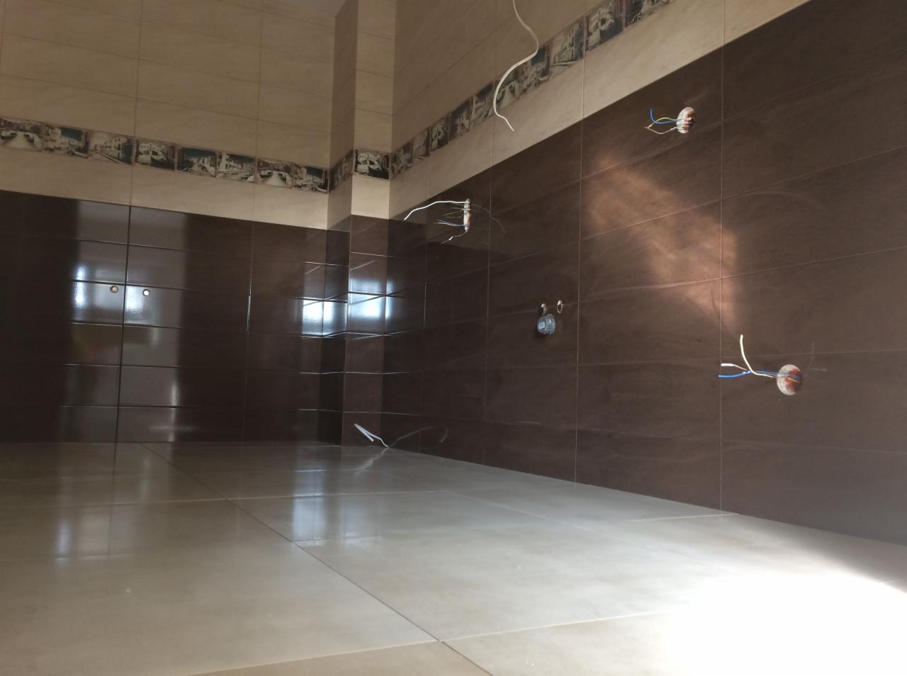 Łazienka w trakcie wykańczania: brązowe płytki ścienne z dekoracyjnym pasem u góry, jasne płytki podłogowe, wystające kable elektryczne ze ścian.