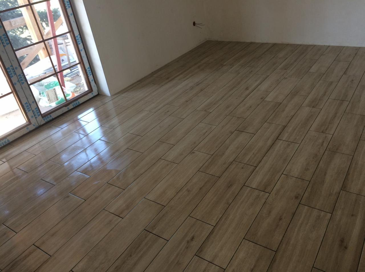 Ułożona podłoga z płytek imitujących drewno w jasnym odcieniu, widok na okno z białymi ramami i fragment ściany z wystającym przewodem elektrycznym.