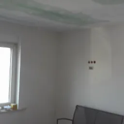 Wnętrze pokoju w trakcie prac wykończeniowych. Widoczne okna, ściany po szpachlowaniu i fragment sufitu z nałożoną siatką i wstępną warstwą gładzi.