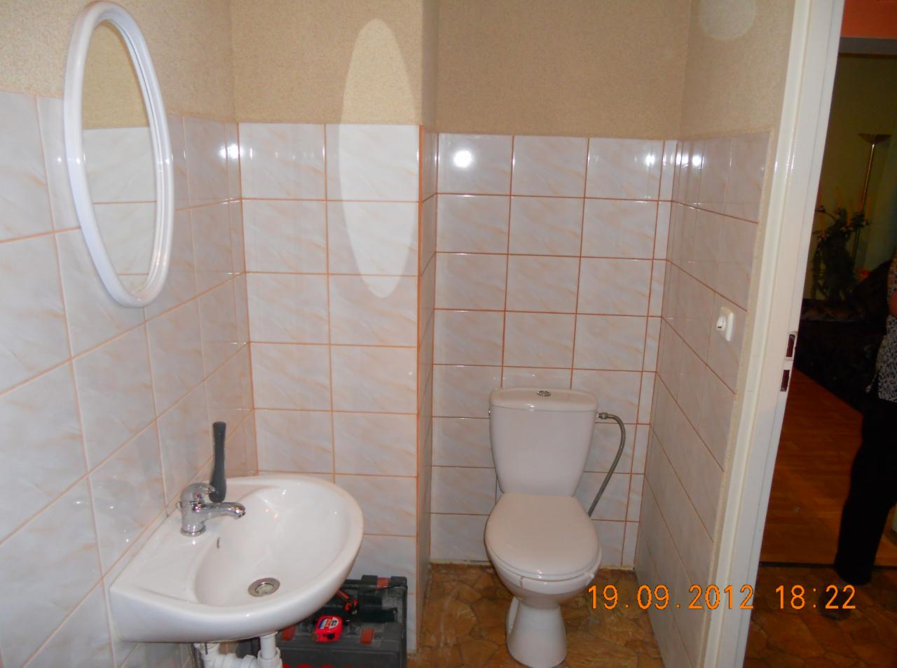 Wykończona łazienka z beżowymi płytkami i pomarańczowymi fugami. Widoczna umywalka, toaleta i lustro. Na podłodze skrzynka z narzędziami.