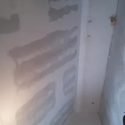 Ściana po szpachlowaniu z widocznymi śladami po łatach, w tle szlifierka do gładzi. Widok z dołu ukazuje etap prac wykończeniowych wnętrza.