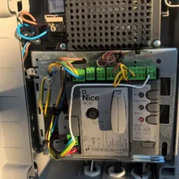Otwarta obudowa sterownika bramy Nice MCA2 z widocznymi przewodami, złączkami i elementami elektroniki. Widoczne oznaczenia i schematy na obudowie.
