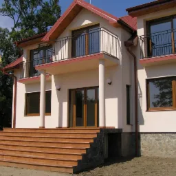 Jasny, dwupiętrowy budynek z balkonami, brązowymi ramami okiennymi i schodami wejściowymi wyłożonymi terakotą, z widocznym podpiwniczeniem wykończonym kamieniem.