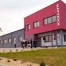 Nowoczesny budynek firmy z czerwoną częścią i logo na fasadzie, widok z zewnątrz, na tle zieleni i szarego nieba. Architektura komercyjna z elementami identyfikacji wizualnej.