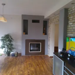 Salon z drewnianą podłogą, kominkiem i ceglaną ścianą. Telewizor wyświetla animację. Wnęki w ścianie z dekoracjami. Nowoczesne oświetlenie sufitowe.