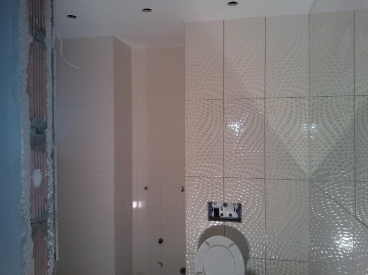 Wykończona łazienka z beżowymi płytkami o wzorze kropek, widoczna toaleta i fragment ściany w stanie surowym. Oświetlenie punktowe w suficie.