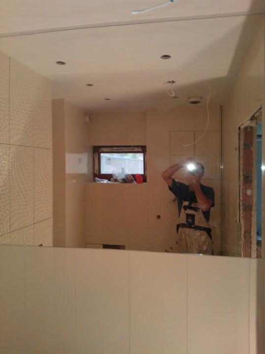 Wykończona łazienka z beżowymi płytkami, widoczna w lustrze, w którym odbija się glazurnik podczas robienia zdjęcia. Okno w tle.