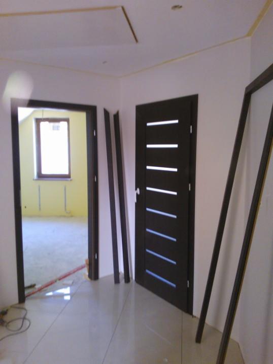 Wnętrze w trakcie wykańczania: widoczne drzwi z poziomymi przeszkleniami, listwy przypodłogowe oparte o ścianę i fragment sufitu podwieszanego.