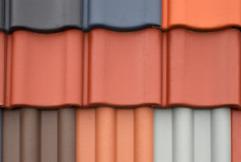 Bliskie ujęcie fragmentu dachu z ułożonymi obok siebie dachówkami w różnych kolorach: szarym, ceglastym, brązowym i białym. Dachówki mają falisty kształt.