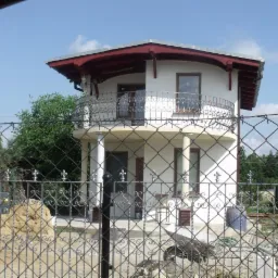 Dwupiętrowy dom z okrągłym balkonem i ozdobną balustradą, widziany zza metalowego ogrodzenia. W tle zieleń drzew i błękitne niebo.