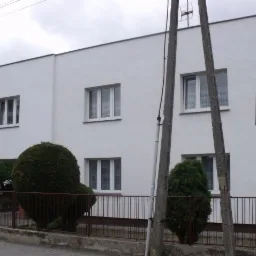 Dwukondygnacyjny, biały budynek z płaskim dachem, widoczny z poziomu ulicy, z elementami architektury modernistycznej. Na pierwszym planie słupy energetyczne i fragment ogrodzenia.