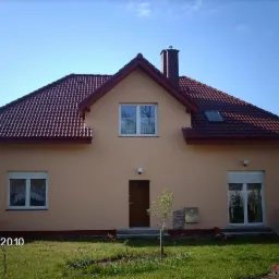 Dwukondygnacyjny dom z beżową elewacją, czerwoną dachówką i niewielkim drzewkiem przed wejściem, sfotografowany w słoneczny dzień.