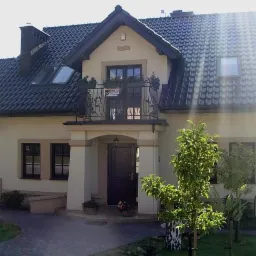 Elegancki, dwukondygnacyjny dom z balkonem i ciemną dachówką. Jasna elewacja, zadbany ogród. Widoczne okna dachowe i numer posesji. Słoneczny dzień.