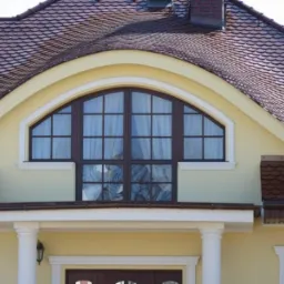 Fragment elewacji domu z brązową dachówką, kremową fasadą i oknem w kształcie łuku. Widoczne kolumny i dekoracyjne elementy architektoniczne.