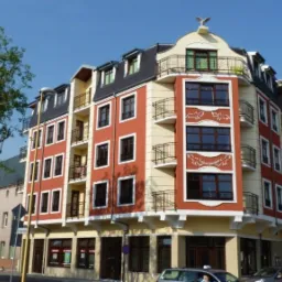 Wielopiętrowy budynek mieszkalny z balkonami i dekoracjami, zwieńczony figurą orła, widziany z poziomu ulicy w słoneczny dzień.