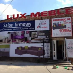 Montera na drabinie montuje baner reklamowy salonu meblowego z ekspozycją mebli pokojowych, młodzieżowych, stołów, krzeseł, sypialni i kuchni na ceglanym budynku, obok widoczna promocja na sofę.