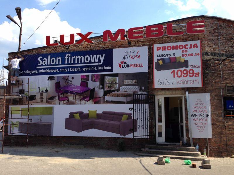 Montera na drabinie montuje baner reklamowy salonu meblowego z ekspozycją mebli pokojowych, młodzieżowych, stołów, krzeseł, sypialni i kuchni na ceglanym budynku, obok widoczna promocja na sofę.