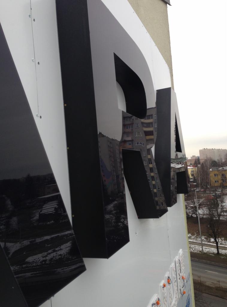 Częściowo zamontowane, czarne litery blokowe z połyskiem, odbijające miejski krajobraz Częstochowy, przymocowane do białej elewacji budynku.