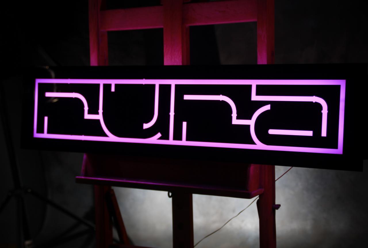 Podświetlany, futurystyczny logotyp z geometrycznych kształtów w kolorze fuksji, umieszczony na czarnym tle i drewnianym stojaku.