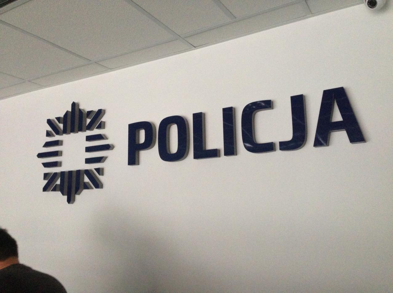 Wykonany z ciemnego tworzywa przestrzenny napis 'POLICJA' oraz symbol na białej ścianie wewnątrz budynku, widoczne z boku i lekko od dołu.