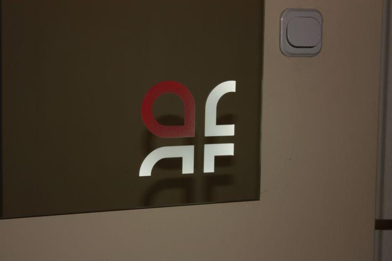 Logo firmy z czerwonym okręgiem i białymi elementami graficznymi na szklanej powierzchni, obok włącznik światła na beżowej ścianie.