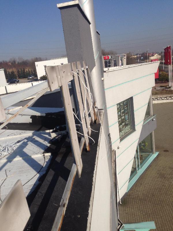 Montaż elementów konstrukcyjnych na dachu budynku, widoczne drewniane podpory i czarna papa, w tle budynek z oknami i masztami flagowymi.