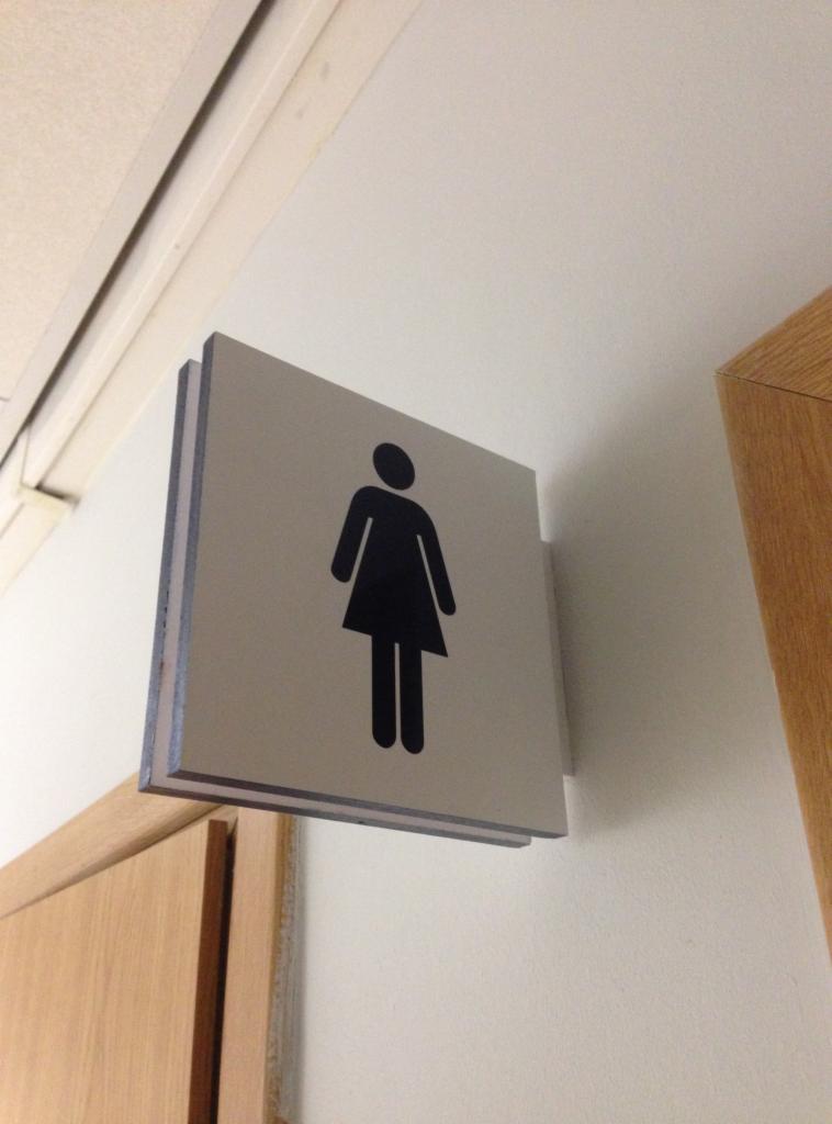 Piktogram kobiety na dwuwarstwowej, kwadratowej tabliczce informacyjnej, zamontowanej na ścianie obok drewnianej framugi drzwi.