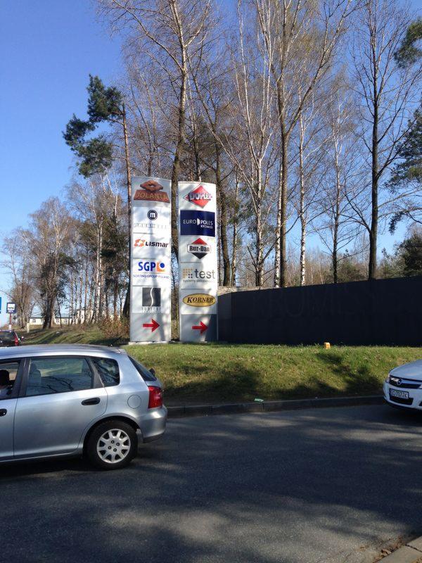 Dwa pylony reklamowe z logo różnych firm, umieszczone przy drodze, widoczne z perspektywy ulicy, z zaparkowanymi samochodami w tle.