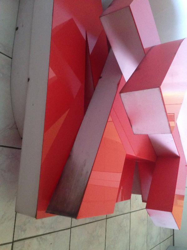 Złożona, przestrzenna litera 'M' w odcieniach czerwieni i różu, wykonana z grubych płyt, zamontowana na kafelkowej ścianie. Widoczne ślady użytkowania na jednej z krawędzi.