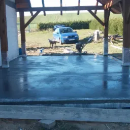 Świeżo wylana, gładka, betonowa powierzchnia pod zadaszeniem, z widoczną zacieraczką, samochodem osobowym z otwartym bagażnikiem w tle i zapasem drewna.